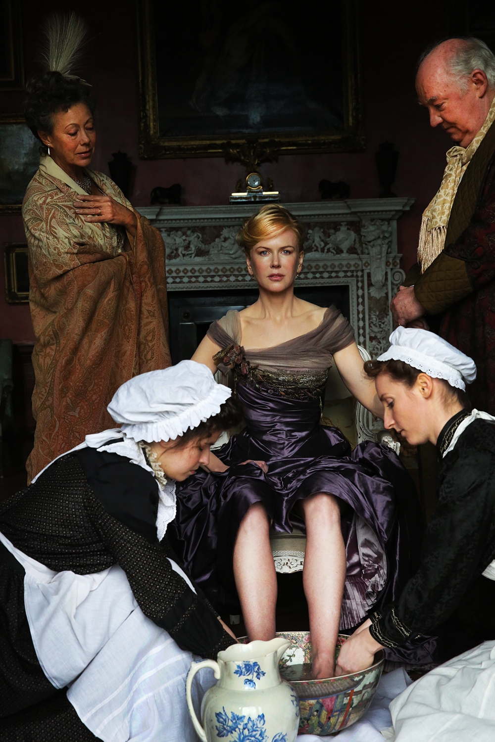 Eine blonde Frau sitzt auf einem verzierten Stuhl und zwei Frauen in schwarz-weißer "Zimmermädchen"-Uniform waschen ihre Füße in einer Porzellanschüssel. Rechts und links stehen ein älterer Mann und eine ältere Frau.