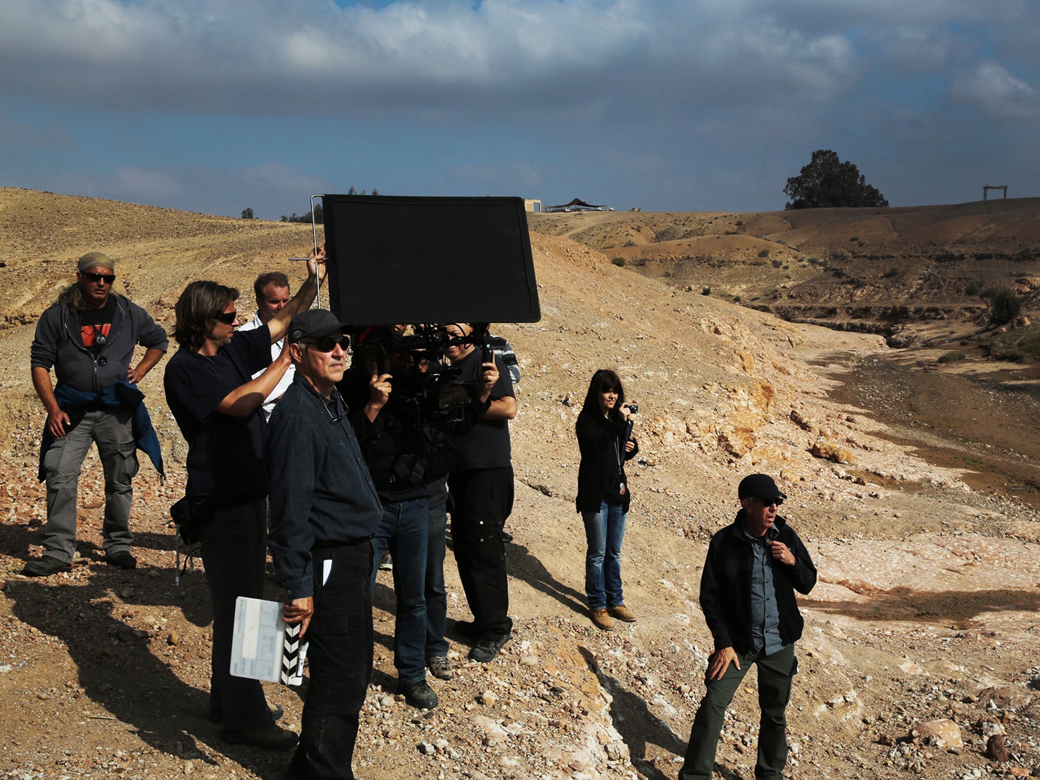 Werner Herzog steht vor seinem Kamerateam und hält eine Regieklappe. Er trägt eine dunkle Kappe, eine Sonnenbrille und dunkle Kleidung.