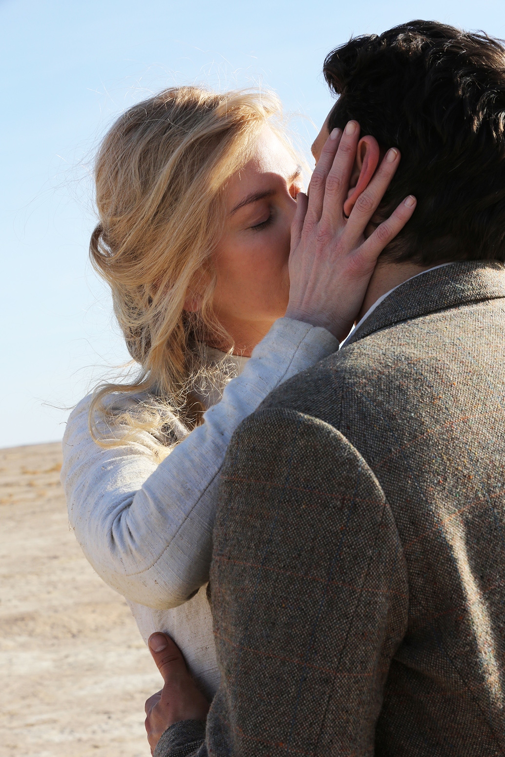 A blonde woman and a dark-haired man are kissing, she holds his face in her hands.