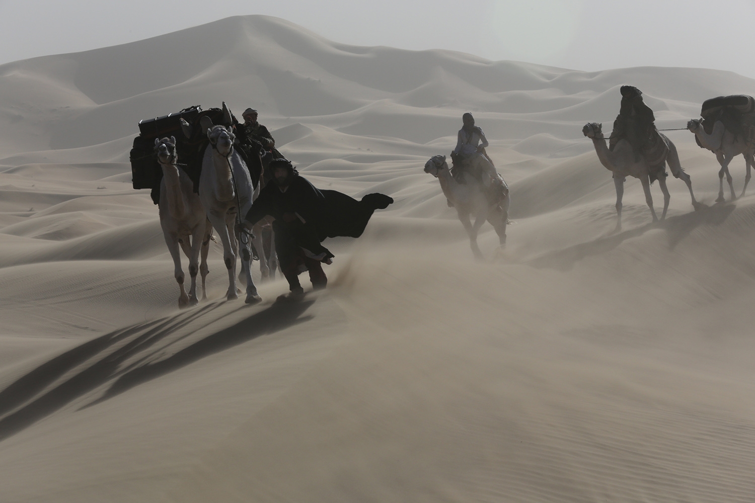 Eine Gruppe Menschen reitet auf Kamelen während eines Sandstrums zwischen Dünen in der Wüste.