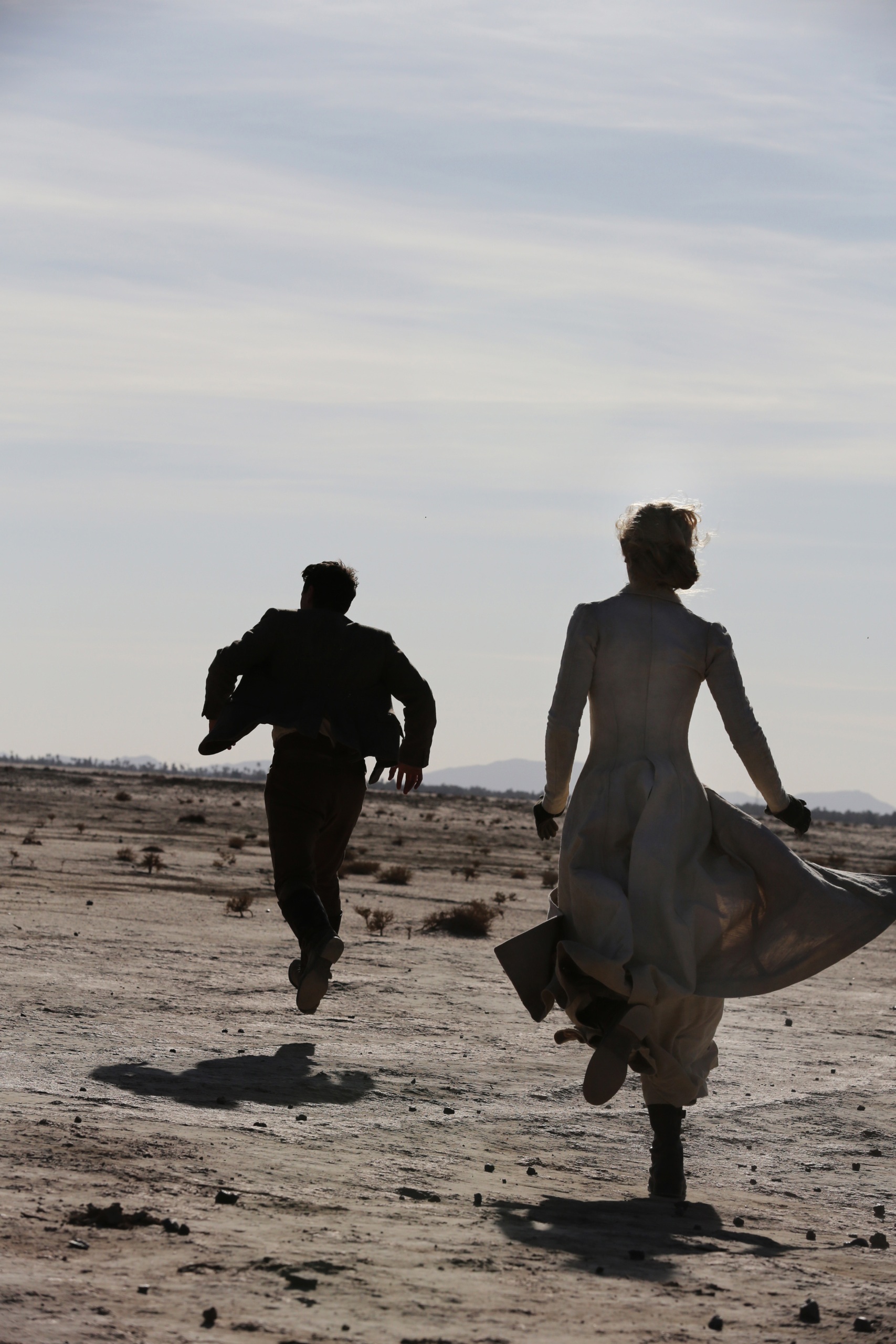 A man in a suit and a woman in a long dress run through the desert, away from the camera. The sun shines intensely and you can only make out the outlines of the people. 
