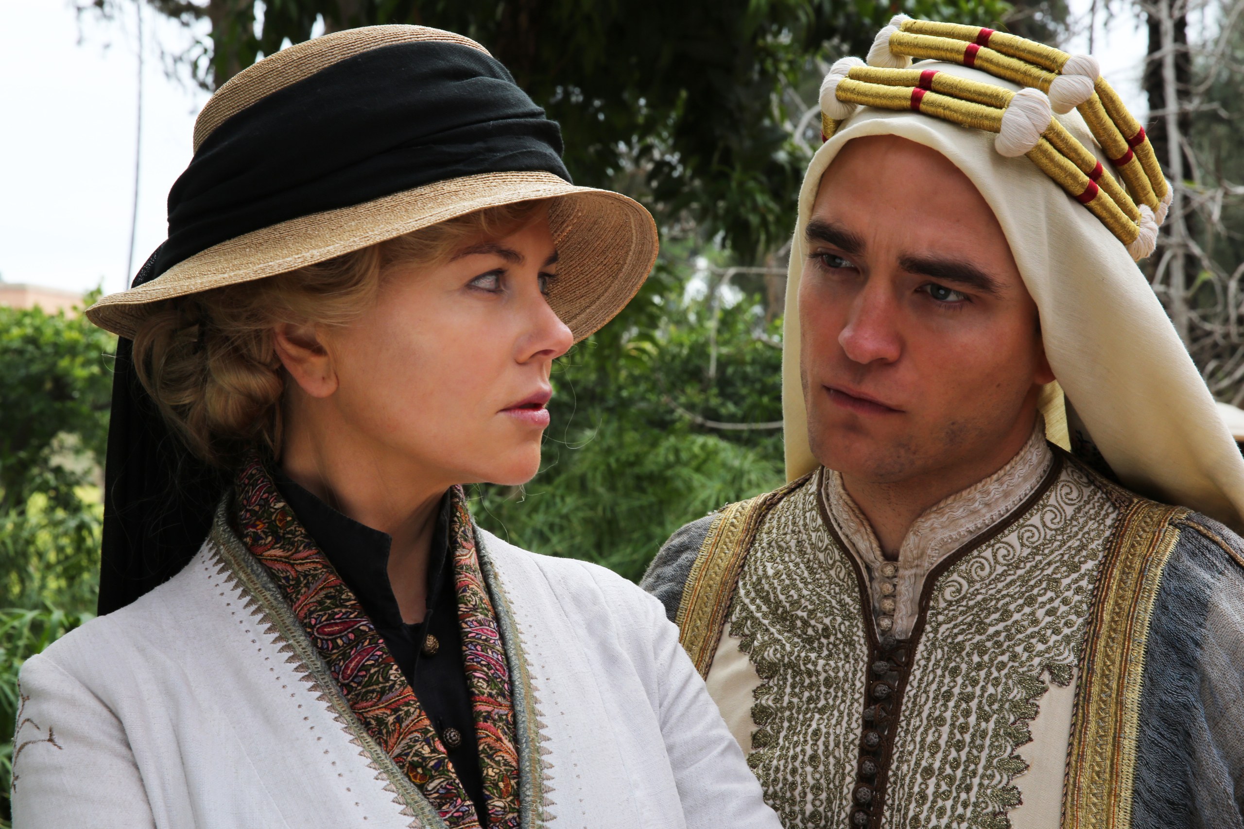 A blond woman wearing a straw hat with a dark ribbon tied around it and a light blouse and a man in an embroidered robe with a head covering look at each other. Behind them you can see green plants.