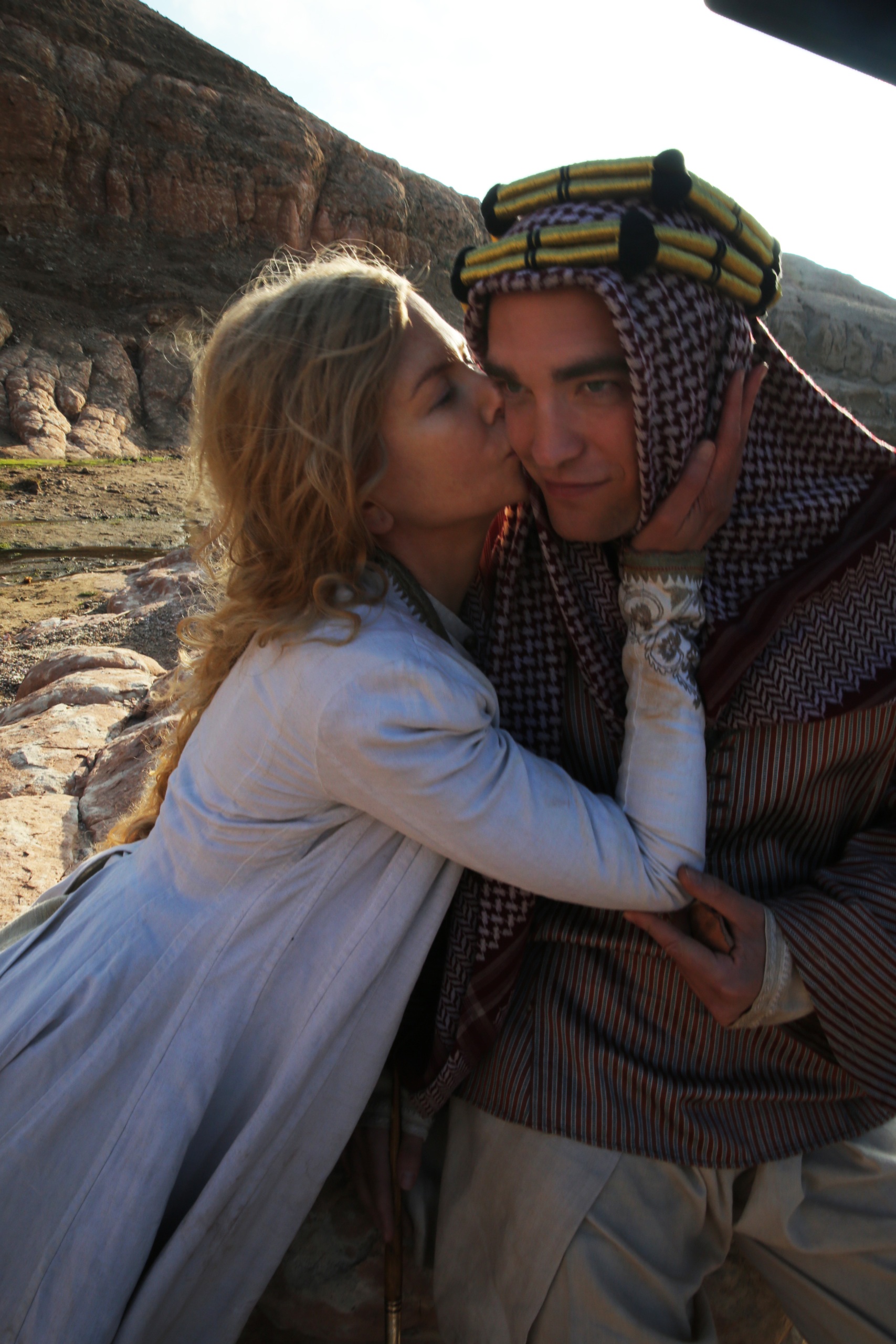 A blonde woman in a white dress kisses a man with a headscarf on the cheek, a rock behind them. 