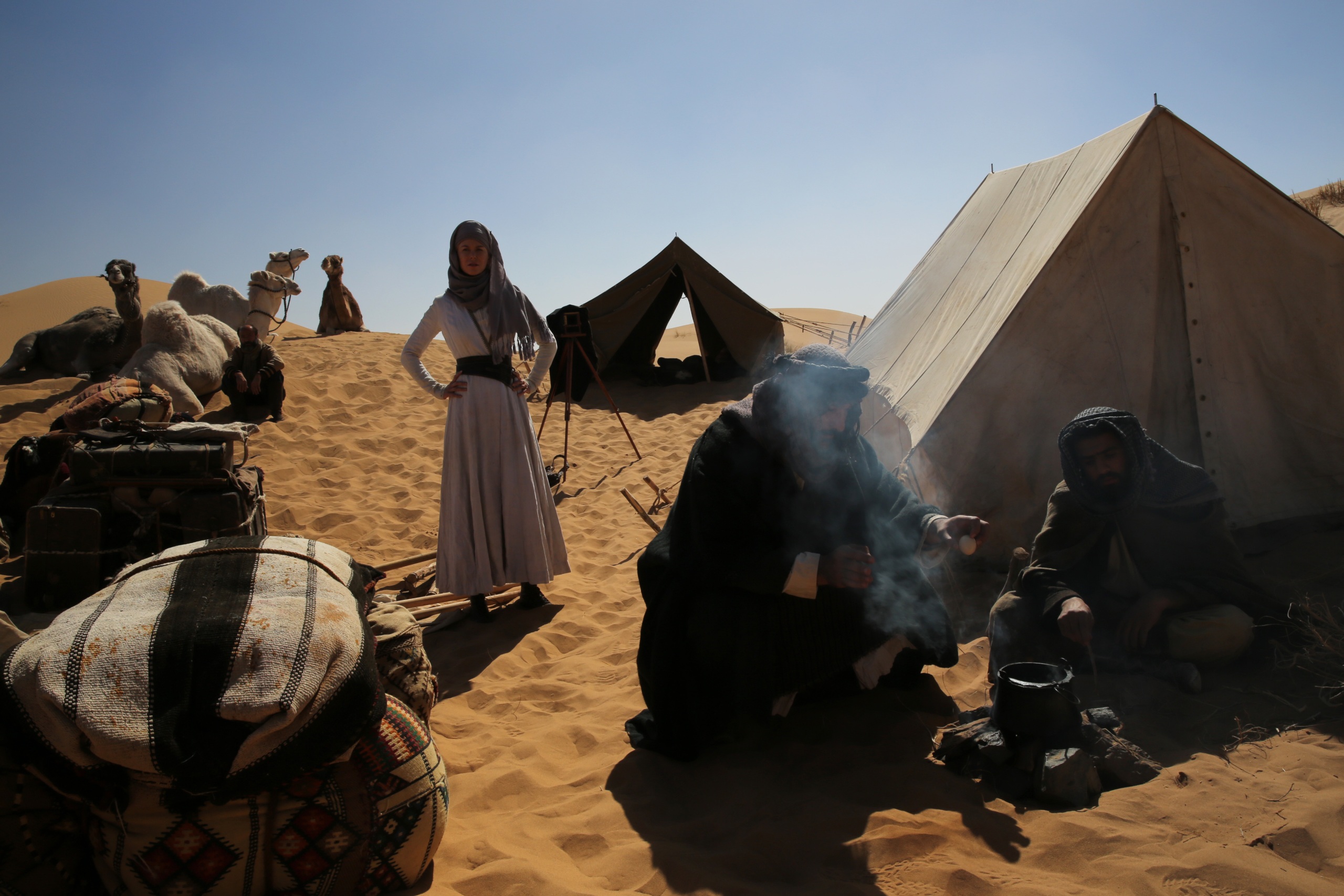 A Bedouin camp in the desert, on the right are two tents, on the left are some camels. In the foreground two people are sitting around a fire, in the background is a woman in a white dress with a dark headscarf.
