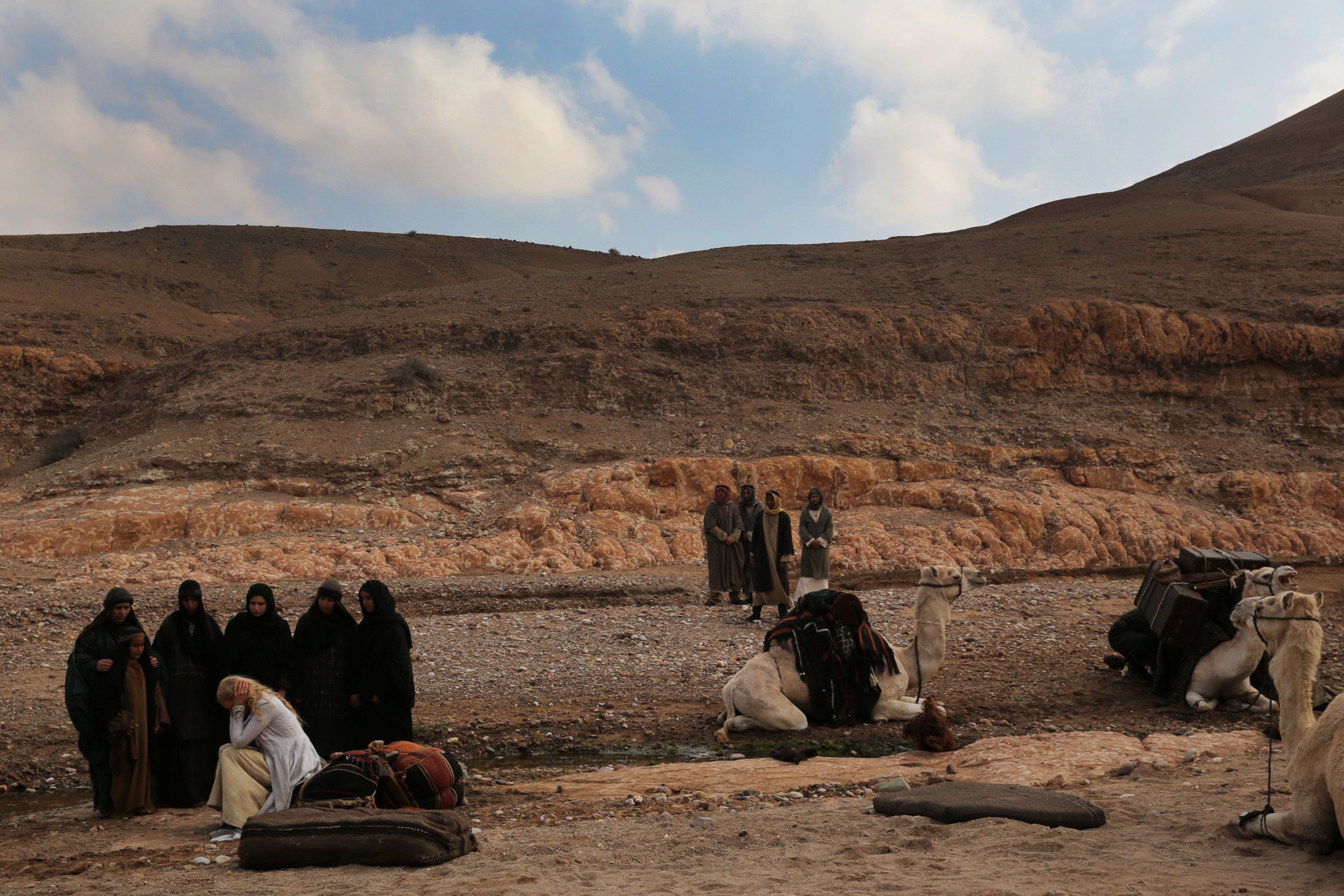 Vor einer Sanddüne in der Wüste liegen rechts drei Kamele, in der Mitte im Hintergrund stehen vier Männer in Kleidung, rechts sitzt eine blonde Frau, das Gesicht in den Händen vergraben und um sie herum stehen sechs weitere Frauen, die sie anschauen. 