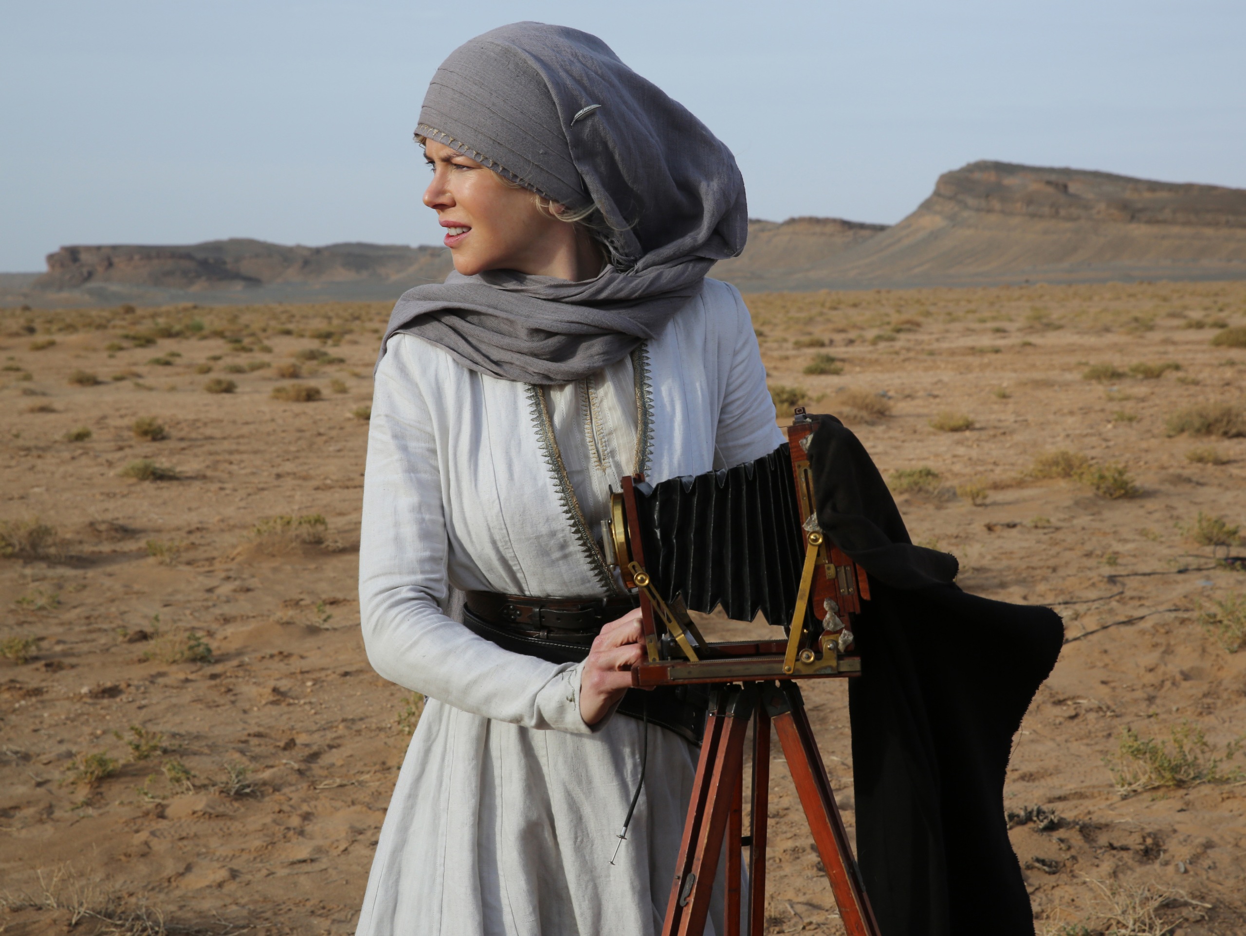 A woman in a white dress with a gray headscarf stands in a desert next to an old photo camera and looks to the left into the distance.