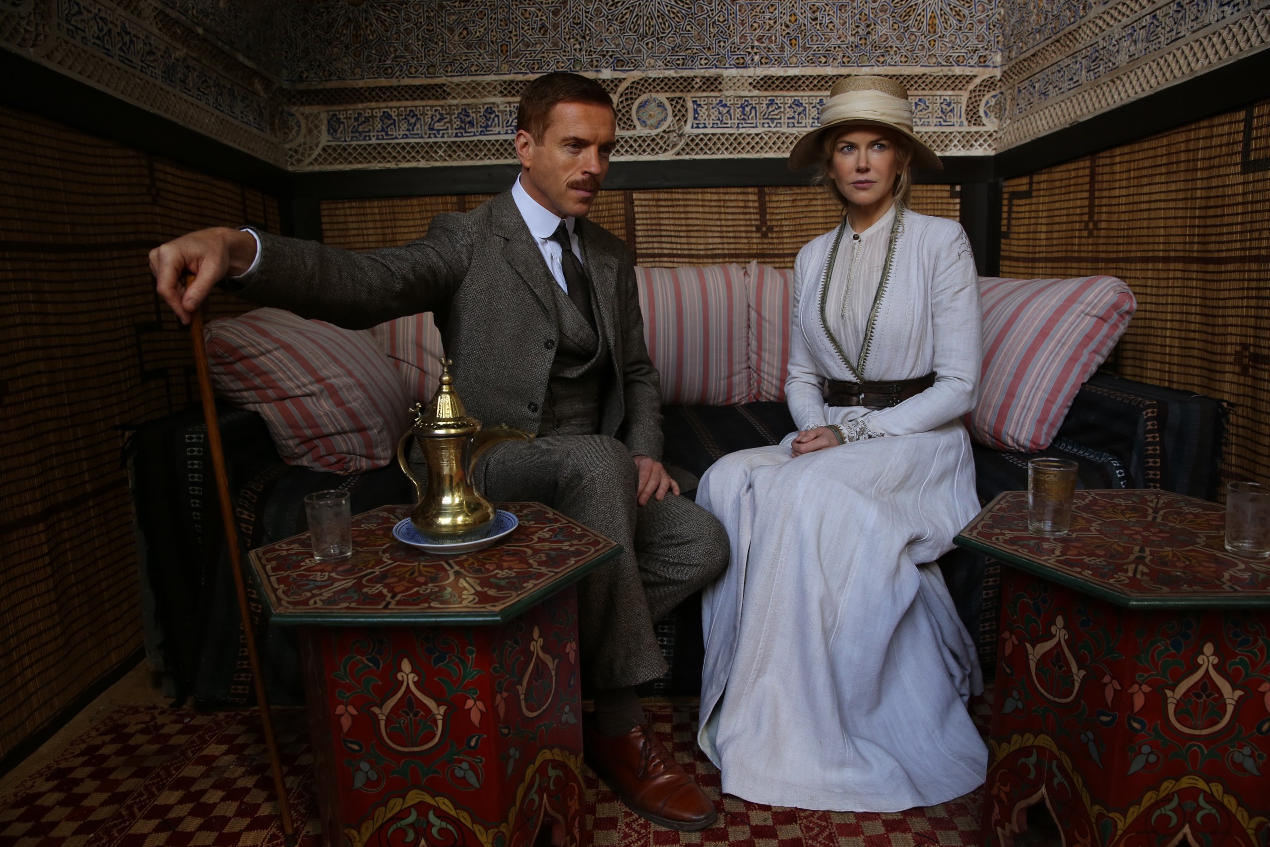 A man and a woman sit side by side on a corner bench in a small room. The woman is wearing a light-colored dress with a straw hat, the man a gray suit, his right hand resting on a stick. In front of them are small, ornate tables with drinks on them.