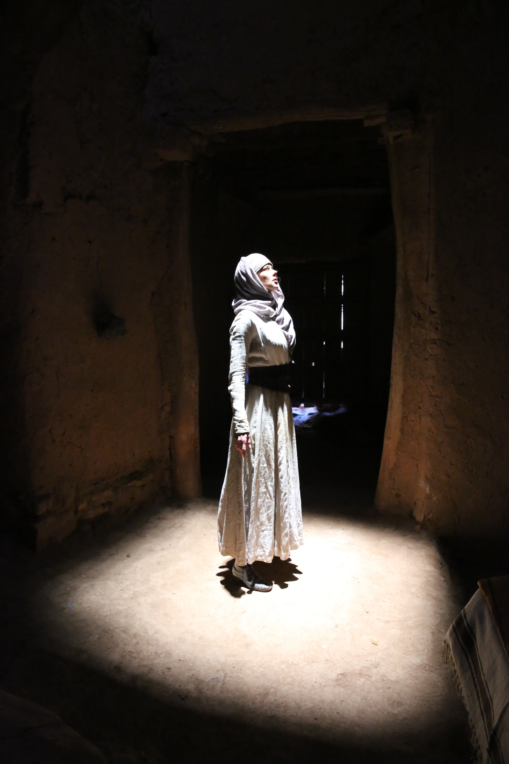 A woman stands in a dark room in the glow of a bright light shining in from above. She wears a light dress and a headscarf.
