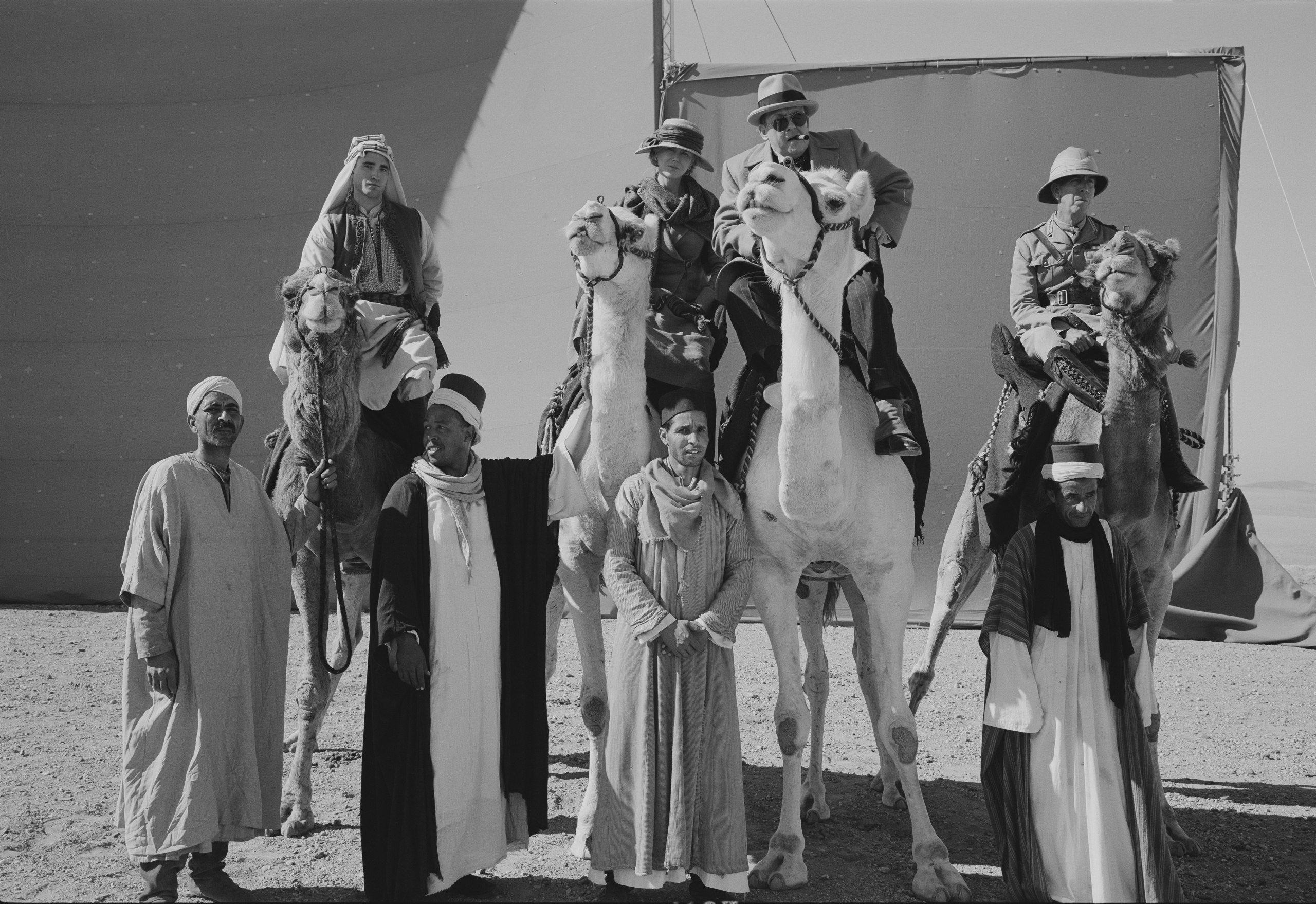 Schwarz-weiß Foto: Vier Menschen sitzen auf Kamelen und vor Ihnen stehen vier Männer, die diese führen.