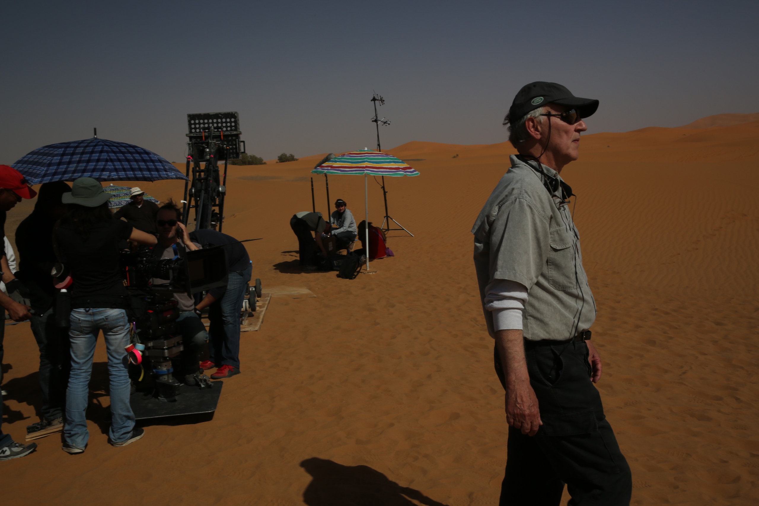 Werner Herzog is standing in the desert, there is a film crew in the background on the right. Herzog is wearing a cap, sunglasses, a light shirt and dark pants. 