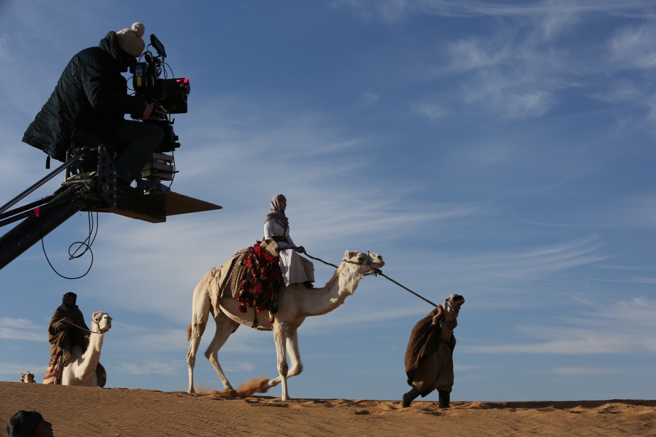 In front of a blue sky in the desert, a woman is led on a camel. Behind her, someone else is riding. On the left in the foreground a man sits on a camera crane and films the described scene.