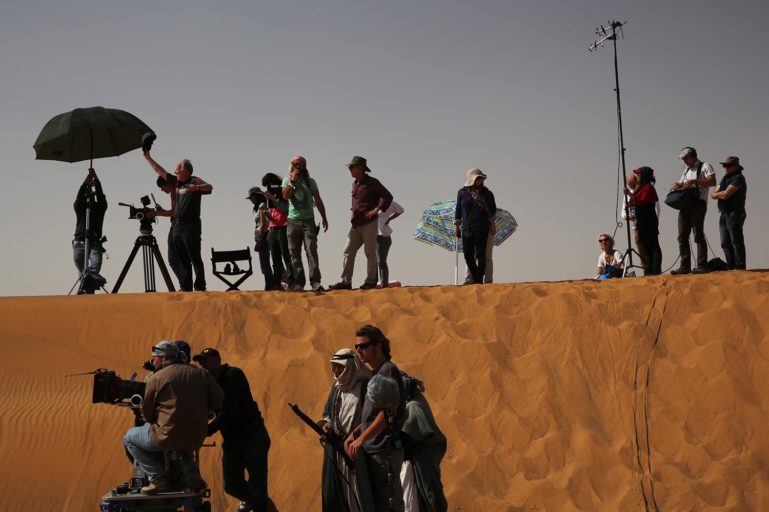 A large group with film equipment stands in the desert, some on a dune, some working with cameras, others talking or observing the situation.