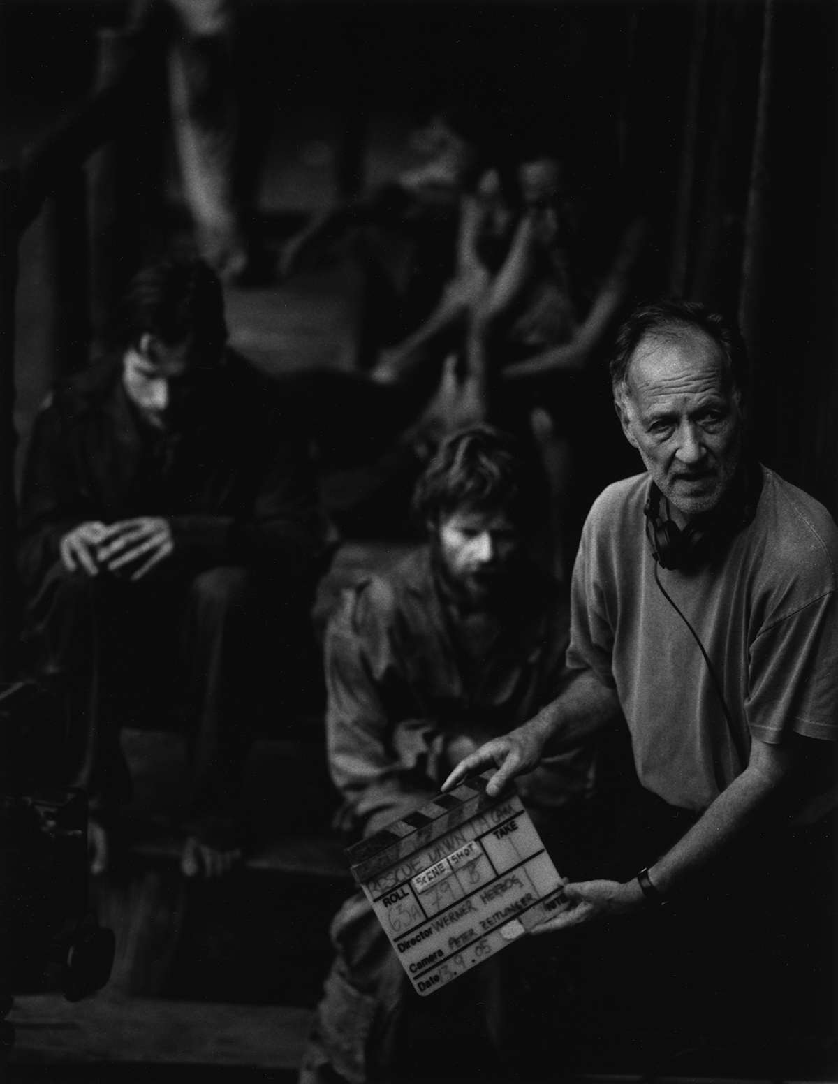 Black and white photo: In the foreground the director with director's hatch, in the background (blurred) the actors are sitting.