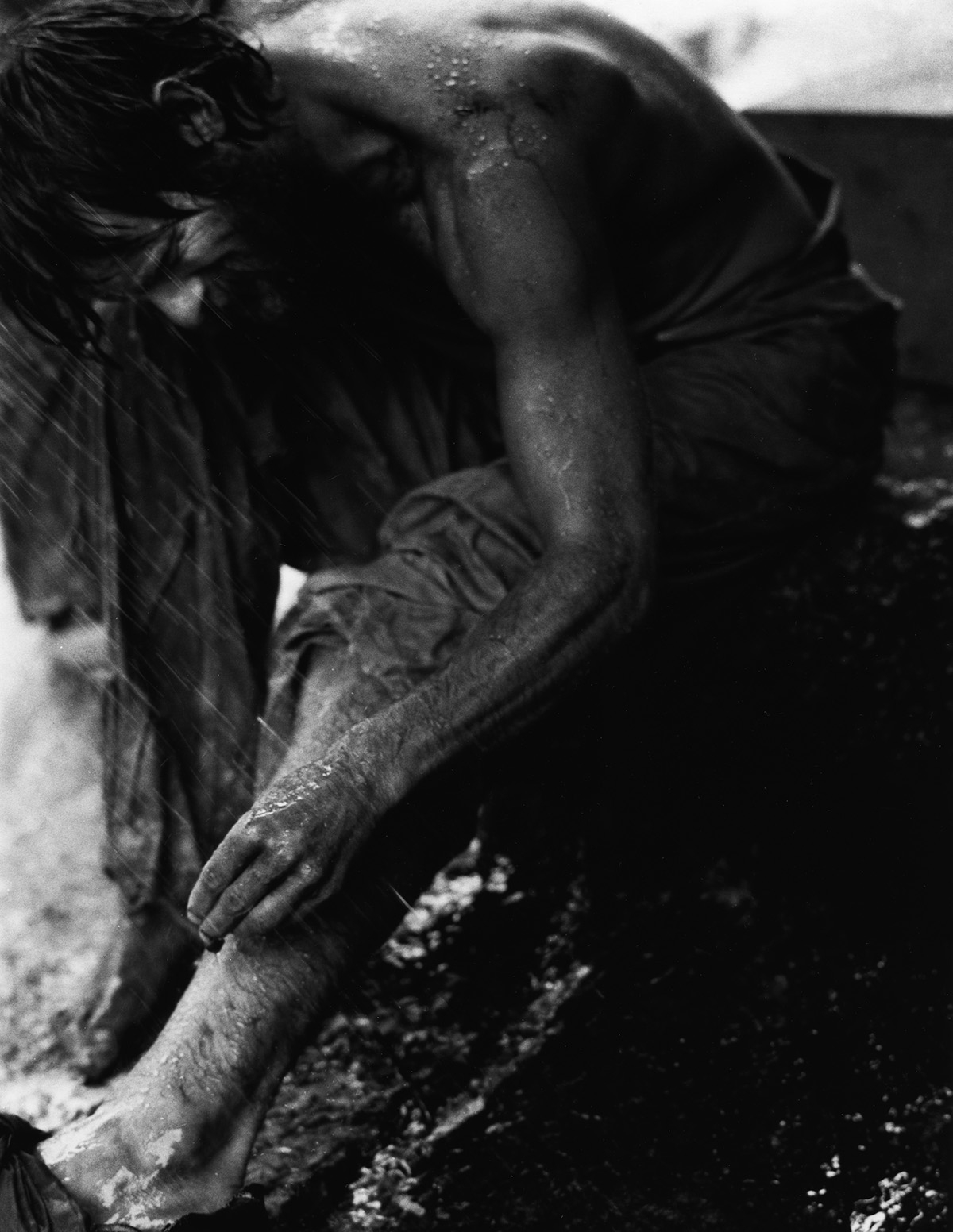 Black and white photo: A bare-chested man in heavy rain, upper body bent forward. He grabs his leg.