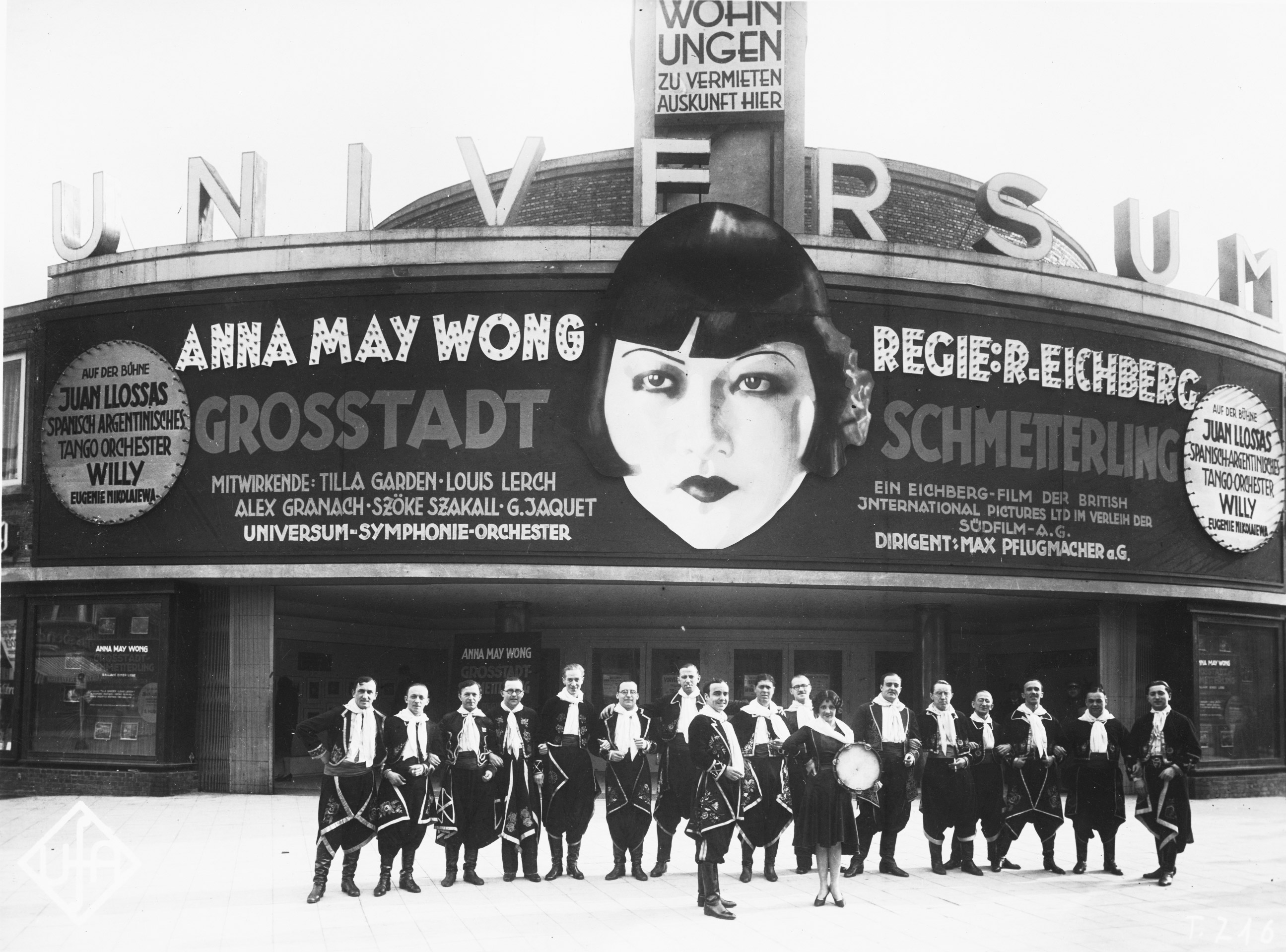 Schwarz-Weiß-Foto: kostümierte Gruppe posiert vor Kinofassade auf der Anna May Wongs Gesicht für einen Film mit ihr wirbt
