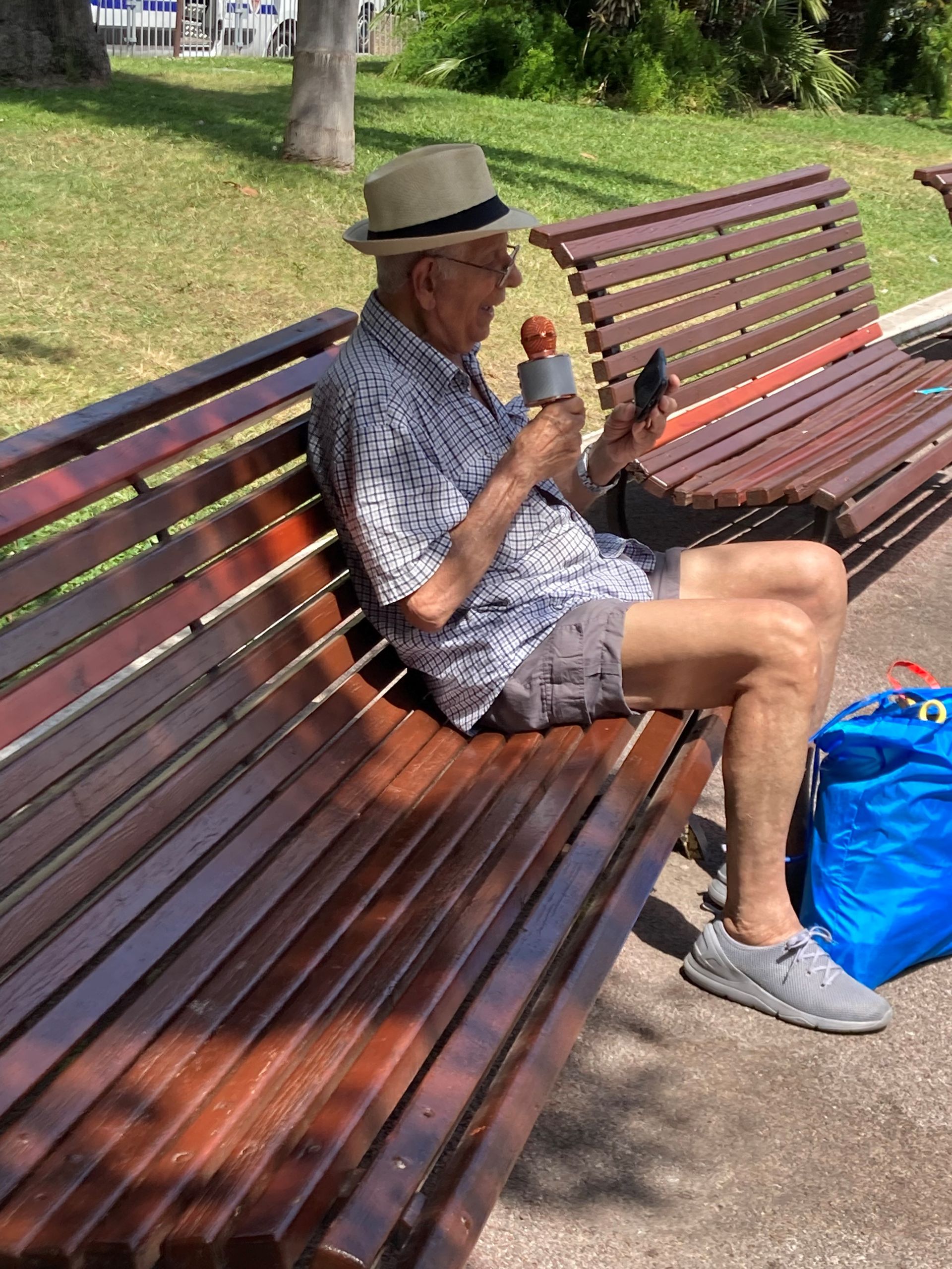 Älterer Herr sitzt auf der Bank, in der rechten Hand ein Handmikrofon, in der linken sein Smartphone