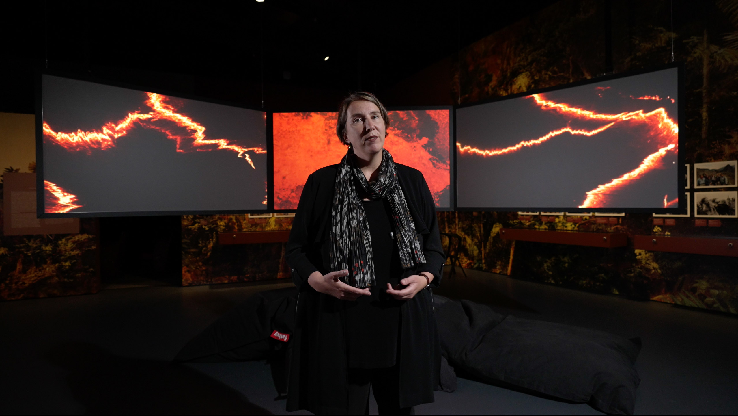 Die Kuratorin der Ausstellung steht vor drei Bildschirmen mit Videos von einem ausbrechenden Vulkan