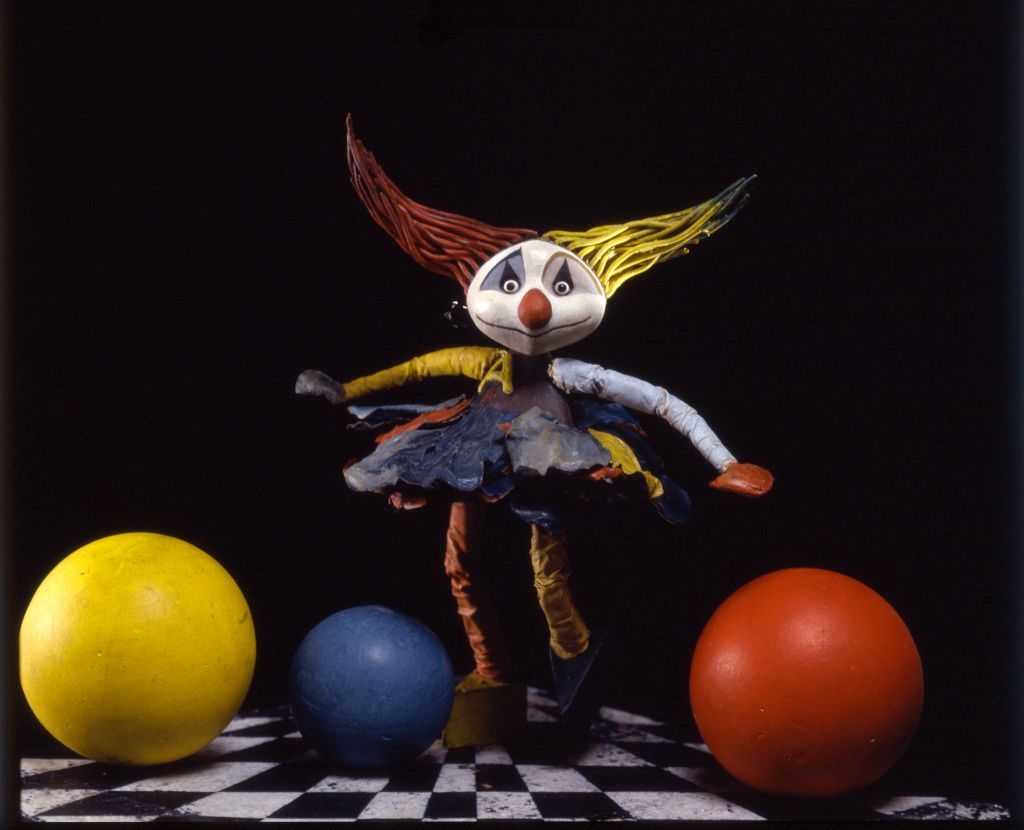 Bunt gekleidetes Mädchen mit Clownsgesicht und gelbroten Haaren steht zwischen drei farbigen Kugeln vor einem schwarzen Hintergrund.