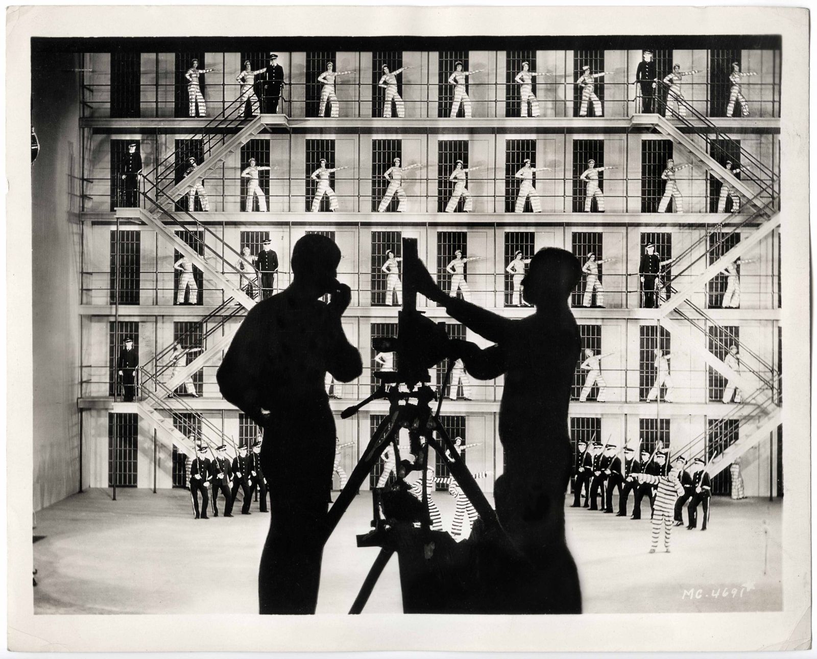 Black and white still from the film Broadway to Hollywood: silhouettes of two persons and a camera in front of a prison set with dancing inmates and guards