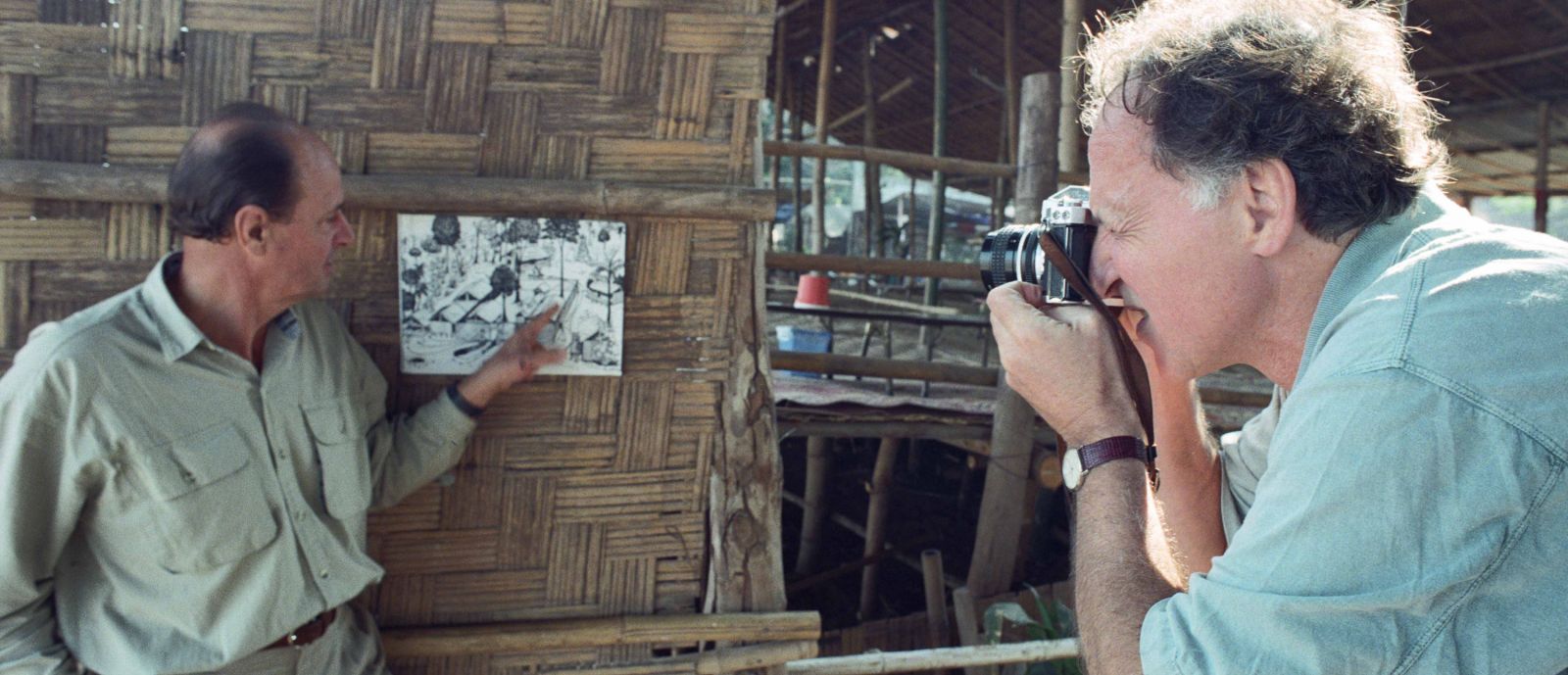 Werner Herzog is taking a photo of Dieter Denger who is pointing at a photograph pinned to a wooden wall.