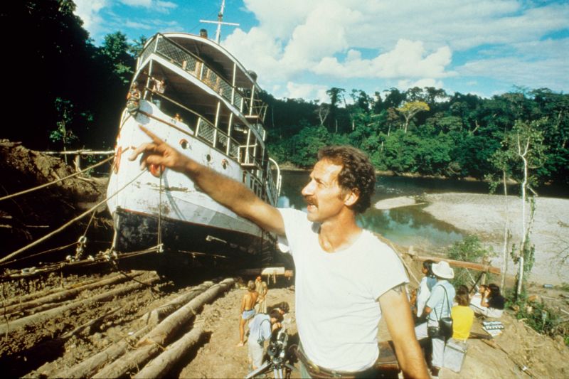 Werkfoto aus dem Film ›Fitzcarraldo‹: Der Regisseur Werner Herzog mit zeigender Geste vor einem Schiff, das auf Land liegt.