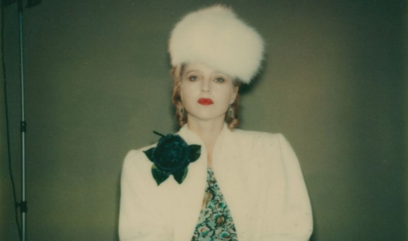 Polaroidfoto einer Frau in weißem Pelzmantel mit einer großen Blume und einem passenden Hut.