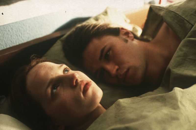 Eine junge Frau liegt mit dem Rücken im Bett, ein junger Mann liegt links von ihr und betrachtet sie.