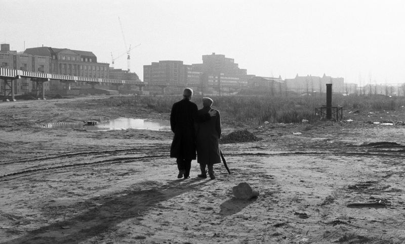 Zwei Menschen in dunklen Mänteln laufen durch eine Berliner Brachlandschaft. Man sieht sie nur von hinten.