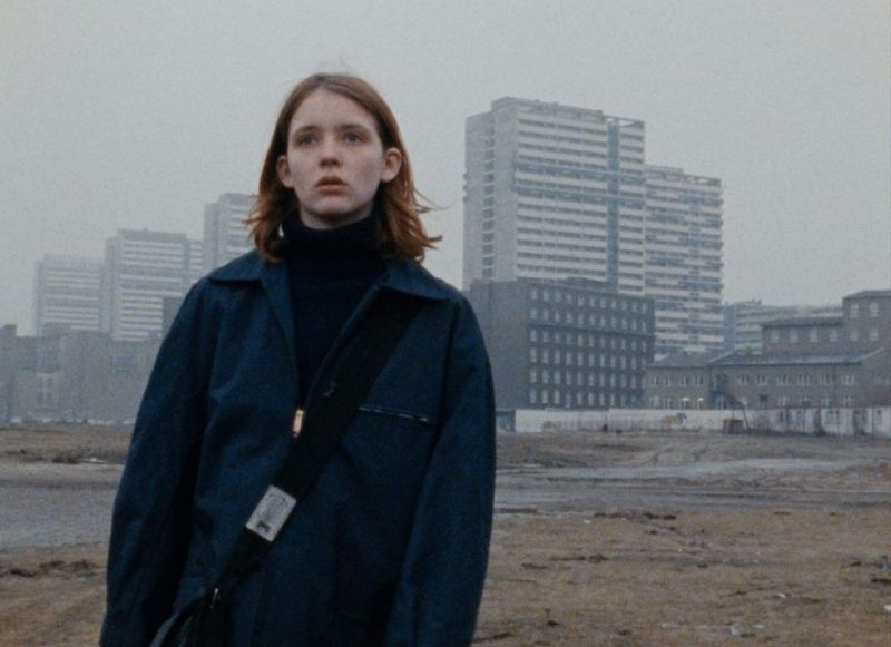 Eine junge Frau steht inmitten einer innerstädtischen Brache, im Hintergrund sieht man Hochhäuser.