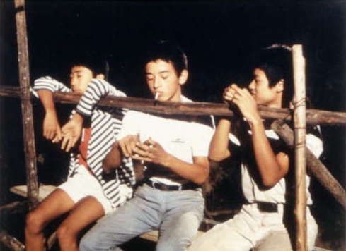 Drei junge Männer sitzen auf einem Baugerüst, der mittlere hat sich eine Zigarette in seinem Mund.