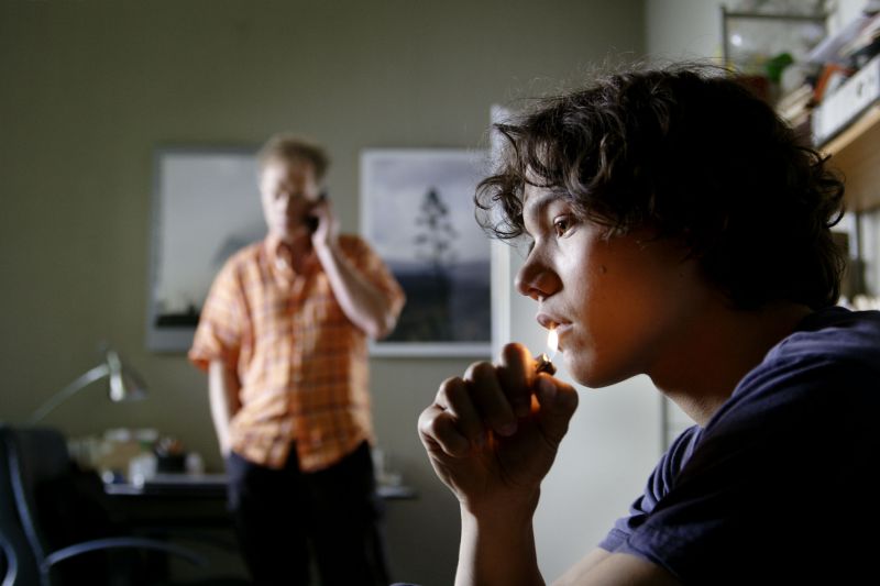 Ein Junge hält ein brennendes Feuerzeug an seine Lippe, im Hintergrund telefoniert ein Mann.
