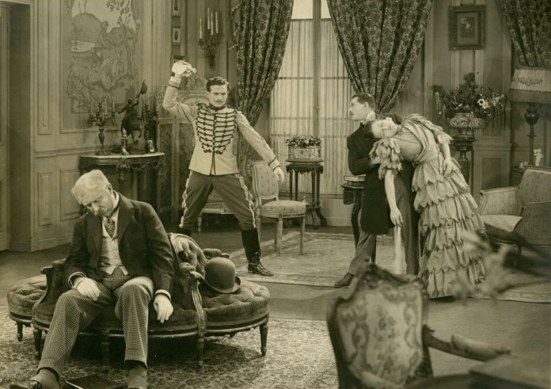 Ein sepia getöntes Bild. Vier Menschen in einem Wohnzimmer des 19. Jahrhunderts. Links döst ein Mann in einem Sessel, hinter ihm schmeißt ein Militär ein Taschentuch auf  den Boden und rechts im Bild hält ein Mann im Anzug eine ohnmächtig gewordene Frau im Kleid.