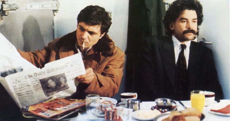 Zwei Männer sitzen nebeneinander am Frühstückstisch; der linke liest Zeitung und raucht, der recht schaut nachdenklich vor sich hin.