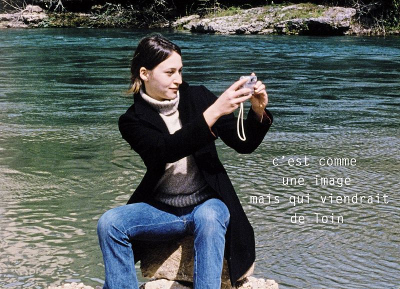 Szenenfoto einer jungen Frau, die vor einem Fluß sitzt und eine Fotokamera in der Hand hält