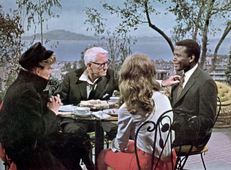 Still in color: Two white women, a white man and a black man sit outside on a patio around a dinner table.nen zum Abendessen gedeckten Tisch. 