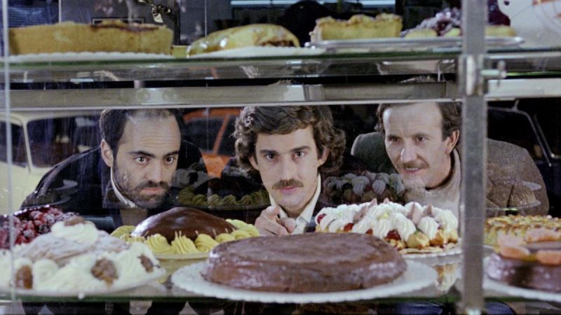 Still in color: Three men are looking through a glass cabinet displaying cakes and the man standing in the middle is pointing at a chocolate cake.