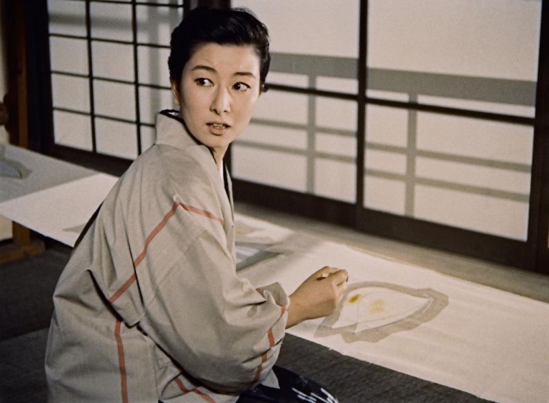 Still in color: A woman kneeling by a window to paint something is looking back into the room over her shoulder.