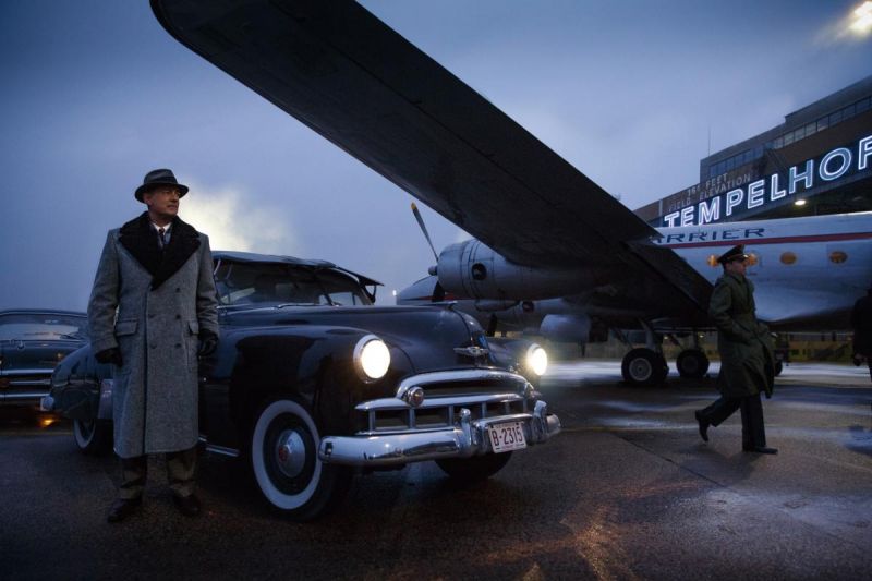Szenenfoto in Farbe: Ein Mann in langem Mantel und Hut steht am Flughafen Tempelhof neben einem Auto unter dem Flügel eines Flugzeuges.