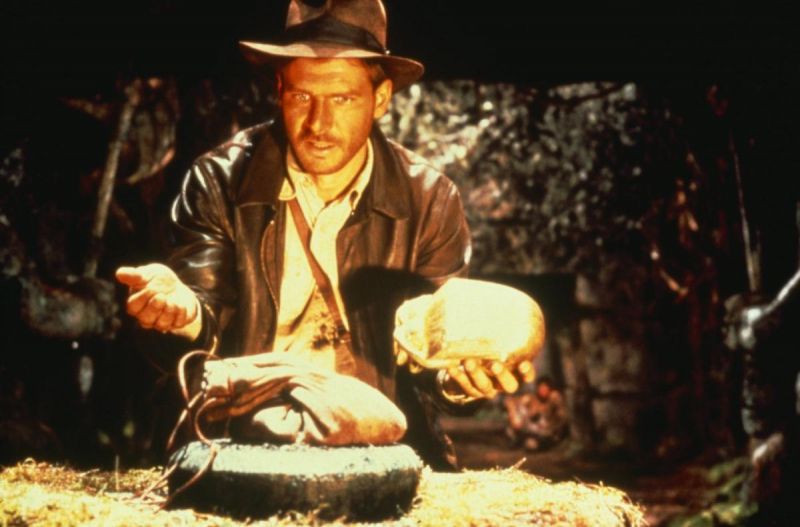 Still in color: A man wearing a cowboy hat and leather jacket is standing in a cave. In front of him a bag is laid out on a bale of straw and he is holding a bundle of fabric in his right hand. 
