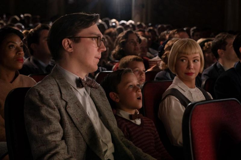 Szenenfoto in Farbe: Ein dunkelhaariger Mann in Anzug, ein dunkelhaariger Junge in Anzug und eine blonde Frau sitzen nebeneinander in einer Sitzreihe wie im Kino.