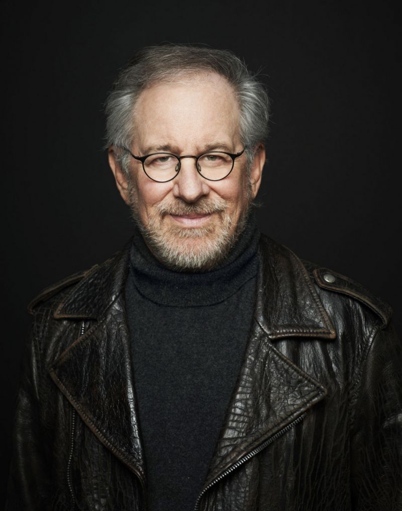 Portrait of Steven Spielberg. He looks friendly into the camera, clothes and background black.