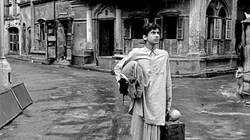 Black and white still: A boy, carrying luggage under his arms, is standing in a derelict street and looking into the distance.