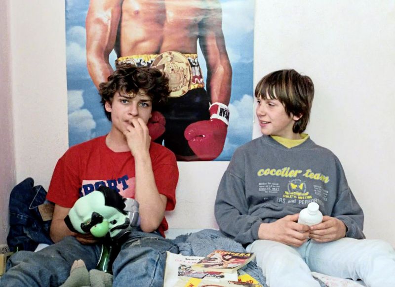 Still in color: Two teenage boys are stitting on a bed infront of a poster showing the torso of a male boxer.