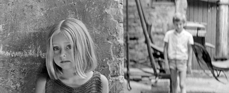 Schwarz-weißes Szenenfoto: Ein blondes Mädchen steht hinter einer Hausecke, rechts hinter ihr steht in einiger Entfernung ein Junge.