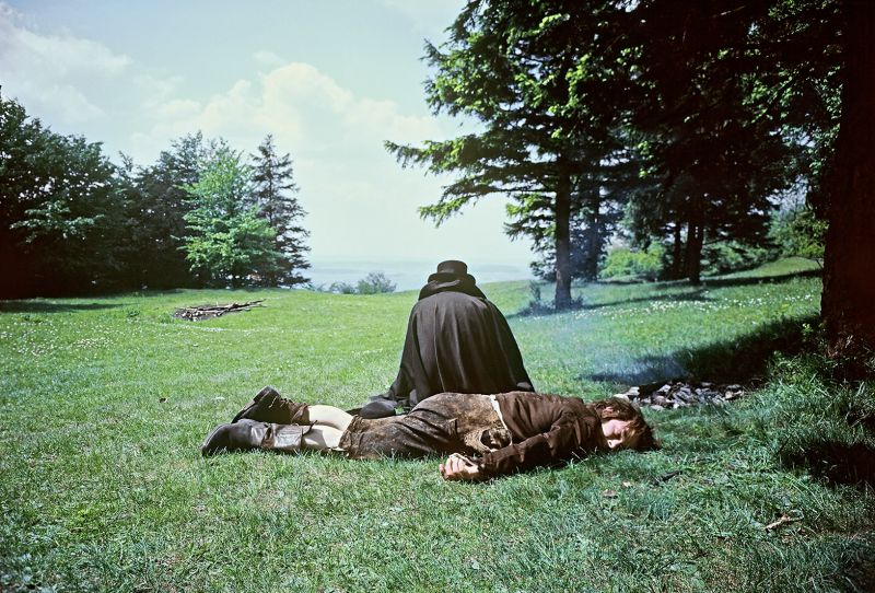 Still in color: On a path in a forrest, a cloaked figure is crouching behind a man lying on the ground.