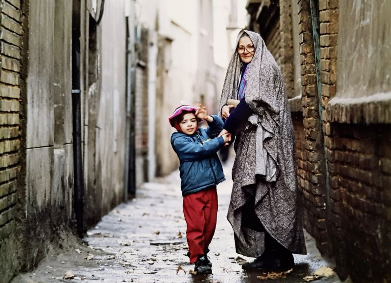 Still in color: A child and a woman are standing in a rainy alley holding hands and facing the camera.