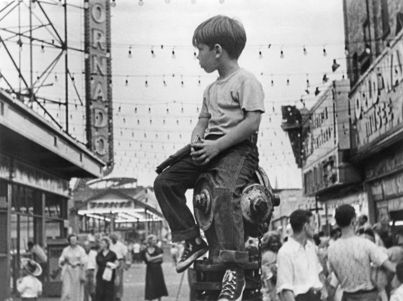 Black and white still: A young boy sitting on a fire hydrant in the middle of an amusement park and watching the people around him.