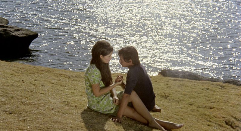 Still in color: A man and woman are sitting closely across from each other on a grassy cliff and looking each other in the eye.
