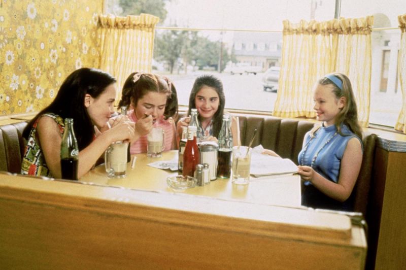 Szenenfoto in Farbe: Vier Teenager-Mädchen sitzen an einem Fenstertisch in einem Diner und trinken Milchshakes.