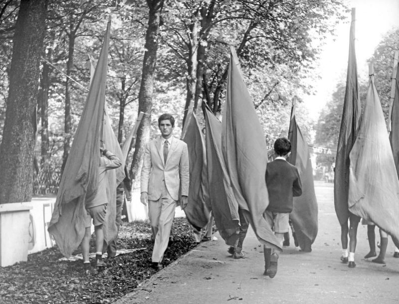 Black and white still: A young man is walking in a park through a group of people holding flags.